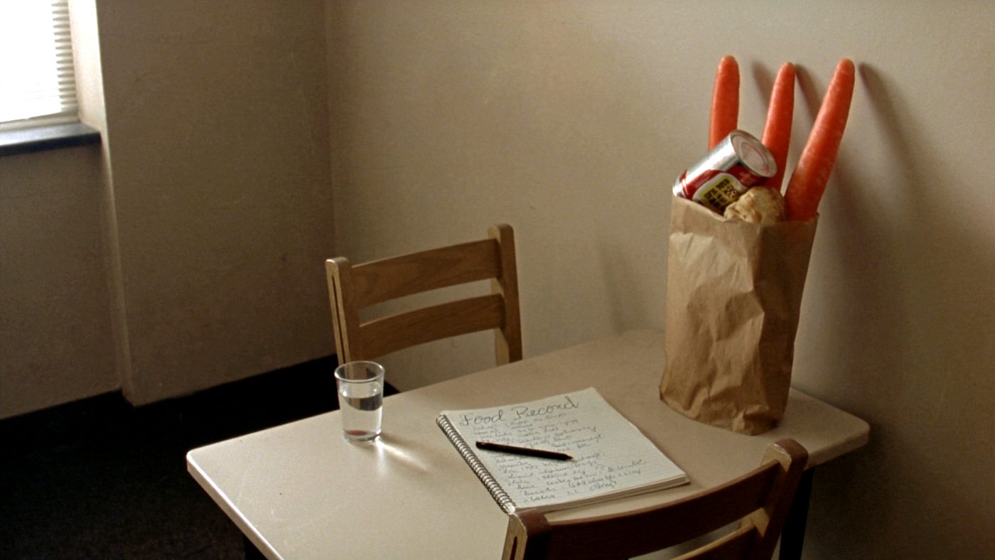 A small plain office table by a window with a spiral notebook headed Food Record, a pen, a glass of water and a paper grocery bag holding carrots, bread and a tin.