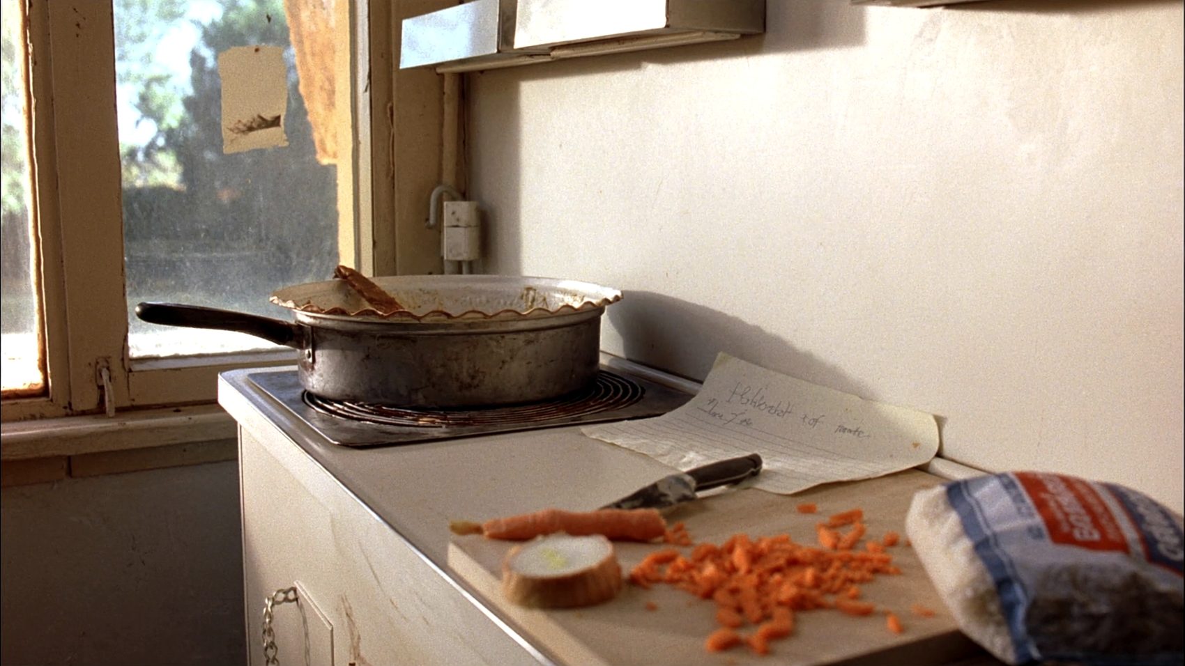 A modest kitchen counter in warm window light: a battered saucepan on an electric hob, a chopping board with chopped carrots and half an onion, a paring knife, a handwritten note on lined paper, and a bag of rice at the edge of the frame.